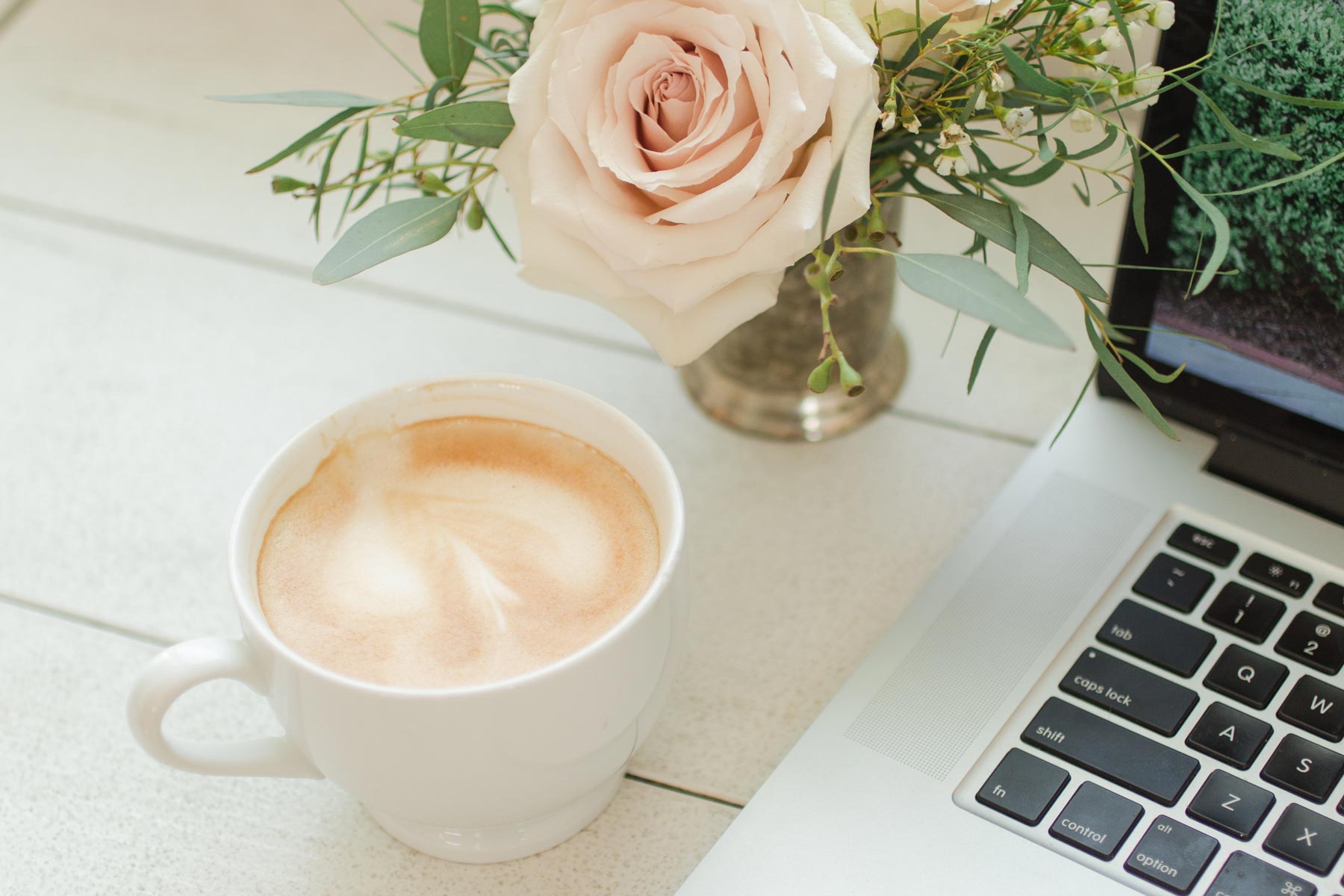 Pink rose on golden vase, coffee cup and blogging laptop on white desk.